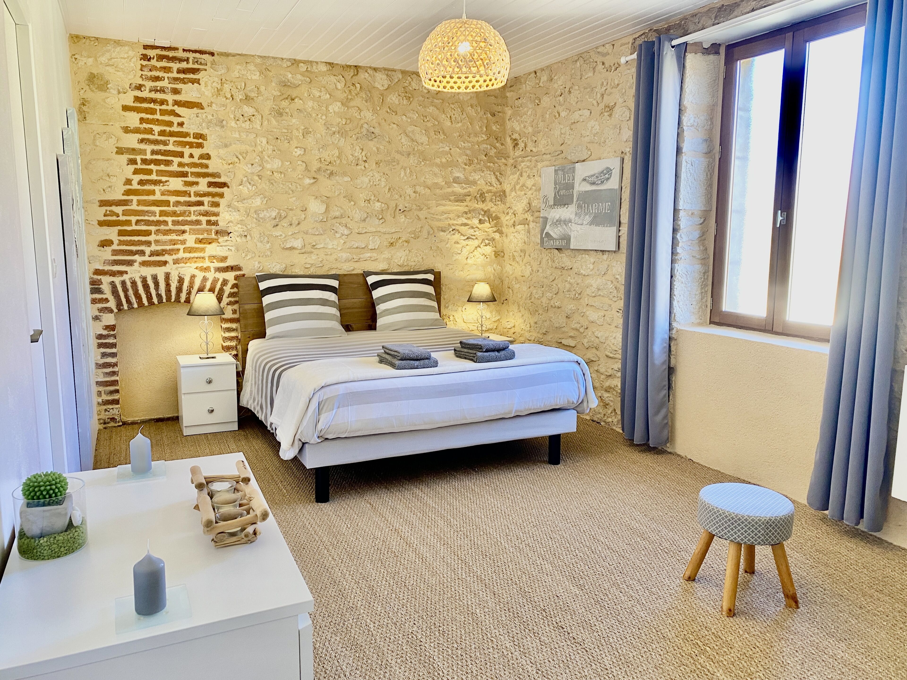 Un dormitorio con pared de piedra, una cama con ropa de cama a rayas, una mesita de noche con lámpara, un taburete pequeño y una ventana con cortinas.