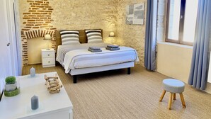 Un dormitorio con pared de piedra, una cama con ropa de cama a rayas, una mesita de noche con lámpara, un taburete pequeño y una ventana con cortinas.