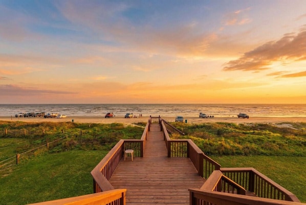 A wooden boardwalk leading to a beach with vehicles and people in the distance.