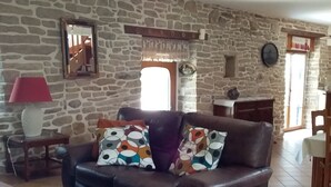 Un salon avec un mur en pierre, un canapé agrémenté de coussins colorés, une petite table avec une lampe et une chaise en bois.