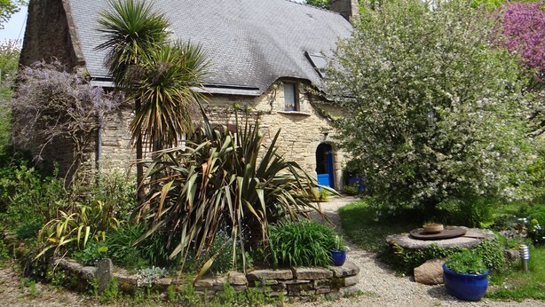 Un bâtiment en pierre, surmonté d’un toit recouvert de tuiles grises, entouré d’une végétation luxuriante et de plantes en fleurs.