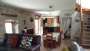 Un salon avec un mur en pierre, un canapé agrémenté d’un coussin coloré, un coin repas avec une table et des chaises, et une cuisine à l’arrière-plan.
