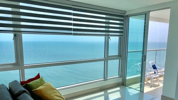 A modern living room with a large glass window overlooking the sea, a grey sofa with colorful cushions, and a balcony with white chairs. - Apartamentos  Cartagena 01 (Cartagena)