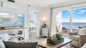 Een moderne woonkamer met een grote televisie waarop een strandscène te zien is, een balkon met uitzicht op de oceaan en een gezellige zithoek.