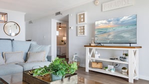 Een woonkamer met een grijze bank, een salontafel met planten, een tv op een houten dressoir, en een muur met ingelijste foto’s en een bord met de tekst 'PARADISE'.