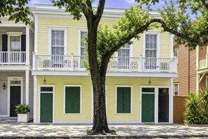 A two-story yellow building with green shutters, a white balcony, and a tree in front.