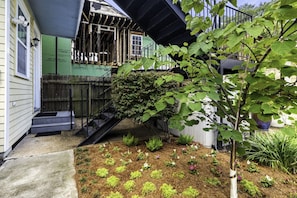 Ein Wohnhaus mit einer Holzterrasse, einem kleinen Garten mit verschiedenen Pflanzen und einer Treppe zu einem überdachten Bereich.