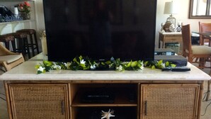 A living room with a flat-screen TV on a marble-top cabinet, wicker furniture, and a decorative garland. - Best Kept Secret on Oahu ! (Waiʻanae)