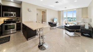 Una cocina moderna con una isla central, un comedor con un taburete de bar, una sala de estar con sofá y televisor, y ventanas amplias con vistas.
