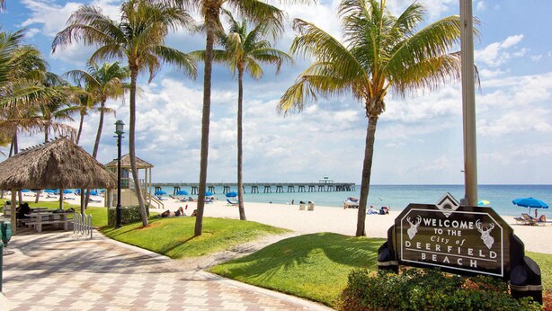 Una playa con un cartel de bienvenida a Deerfield Beach, un muelle y palmeras.