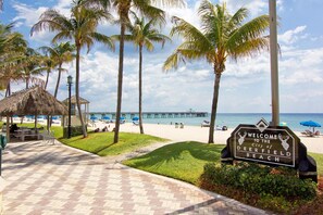 A beach with a sign welcoming visitors to Deerfield Beach, a pier in the background, and a thatched-roof structure.
