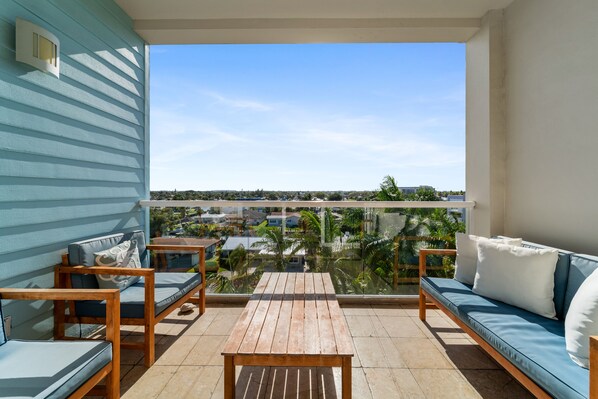 A balcony with a wooden table and two sofas, offering a view of a residential area with houses and palm trees.