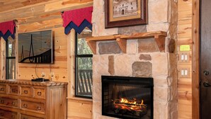 Un acogedor interior de cabaña con chimenea de piedra, armarios de madera y un televisor montado.