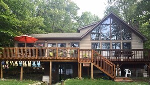 A two-story building with a wooden deck and a covered area with an umbrella. - Windy Break Lake Front House (Gouldsboro)