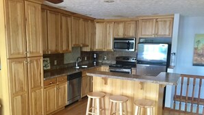 A kitchen with wooden cabinets, a stainless steel refrigerator, a microwave, a stove, and a dishwasher. There are three wooden stools at the counter. - Windy Break Lake Front House (Gouldsboro)
