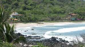 Une plage au rivage rocheux, avec quelques maisons et une forêt dense en arrière-plan.