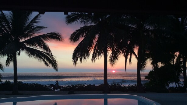 Une vue sur le coucher de soleil depuis un espace aménagé près de la piscine, avec des palmiers et un ciel dégagé.