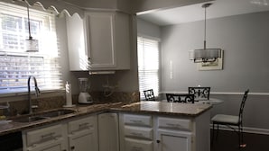 A kitchen with white cabinets, a granite countertop, a built-in dishwasher, and a dining area with a table and chairs. - Carmel Road / Ballantyne  / South Charlotte Area Cape Cod  Home (Charlotte)