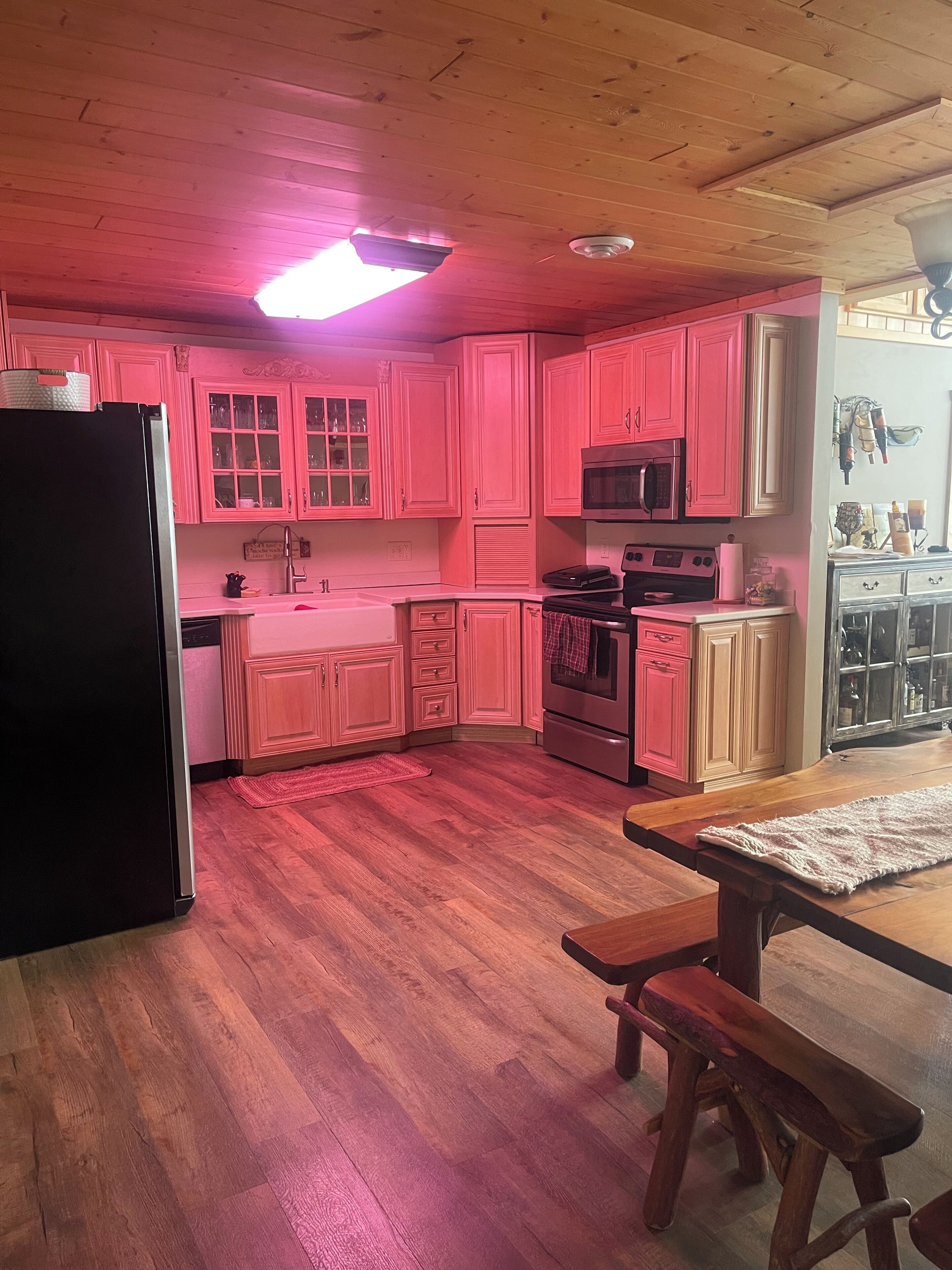 Una cocina con armarios rosados, un refrigerador negro, un microondas y una mesa de madera con un banco.