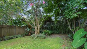 A backyard with a wooden fence, a tree with pink flowers, and various plants. - Large Shotgun Double In Perfect Uptown Location Near Magazine and State St. (New Orleans)