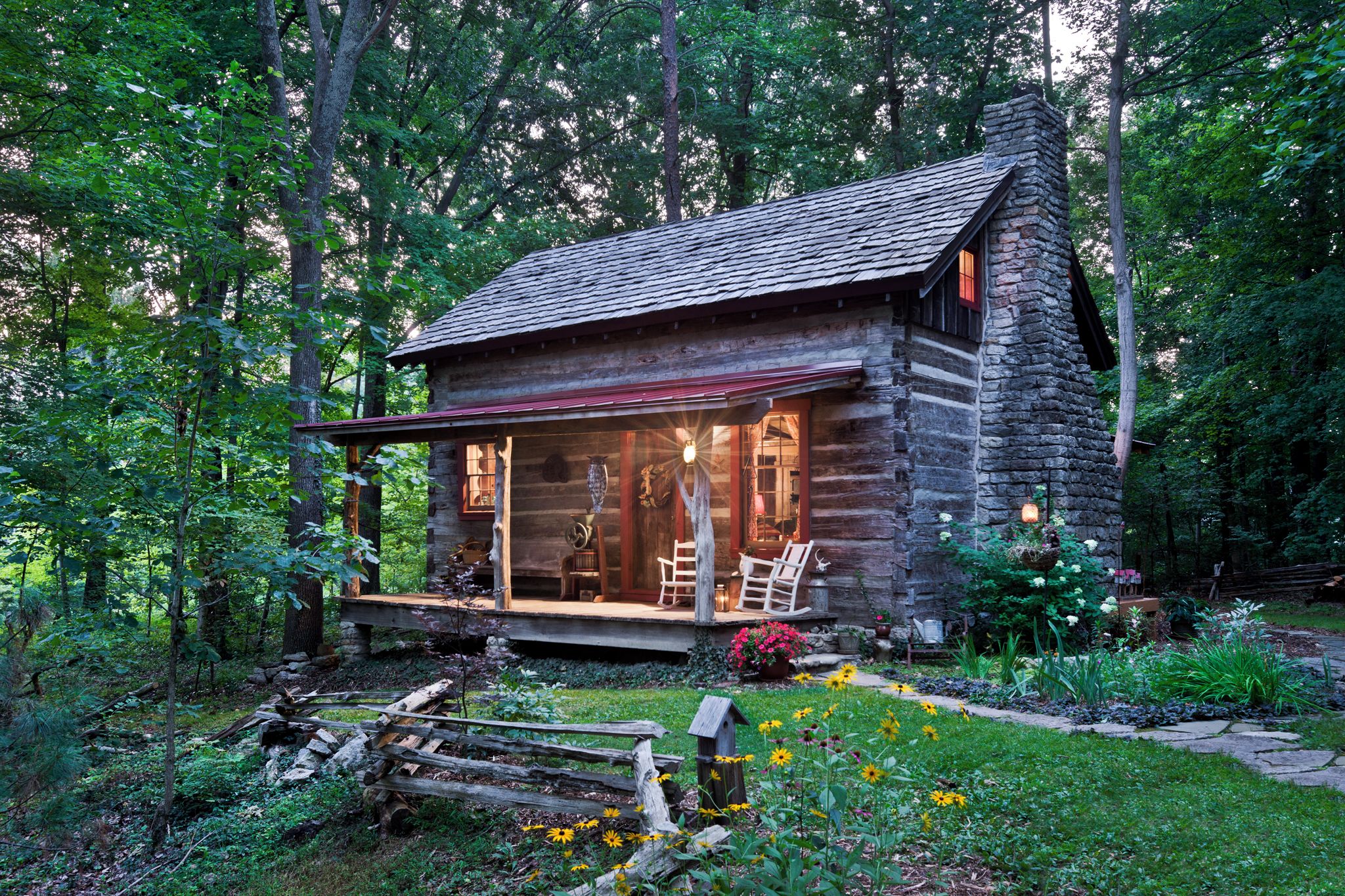 A rustic cabin with a stone chimney, wooden porch, and a garden with flowers and a wooden fence.