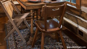 Interior rústico de una cabaña de madera con una mesa de comedor, sillas, una araña y una ventana con cortinas.