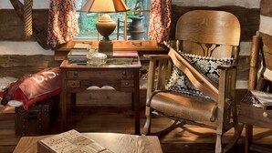 Interior rústico de una cabaña con un escritorio de madera, una mecedora y una ventana con cortinas.