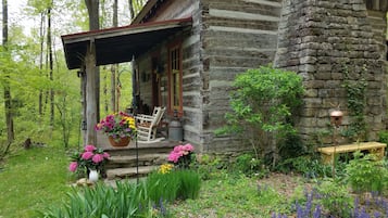 Un chalet rustique en pierre, doté d’un porche, entouré d’une végétation luxuriante et de plantes en fleurs.