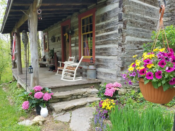 Une cabane rustique en rondins de bois, dotée d’un porche, d’une chaise berçante et d’un panier à fleurs suspendu.
