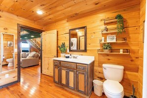 L’intérieur d’une cabane en bois est doté d’un plancher en bois, d’un évier encastré avec un miroir, de toilettes, d’un canapé et d’un escalier menant à une autre pièce.