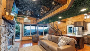 A spacious living area with a stone fireplace, a leather sofa, and a kitchen with modern appliances. - Big Time Hill Cabin with an indoor pool, hot tub, & great views! (Bryant)