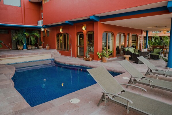 A swimming pool with lounge chairs, surrounded by potted plants and a building with arched windows.