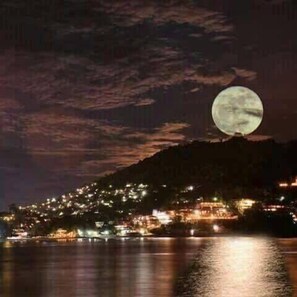 A night scene with a full moon over a coastal town with illuminated buildings.