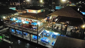 Un ristorante all'aperto su più livelli, con tavoli e ombrelloni, circondato da un pontile in legno e una struttura con tetto di paglia.