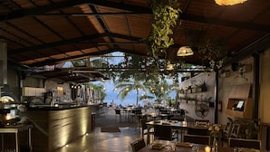 En restaurang inomhus med utsikt över stranden, med en bardel, matbord och hängande växter.