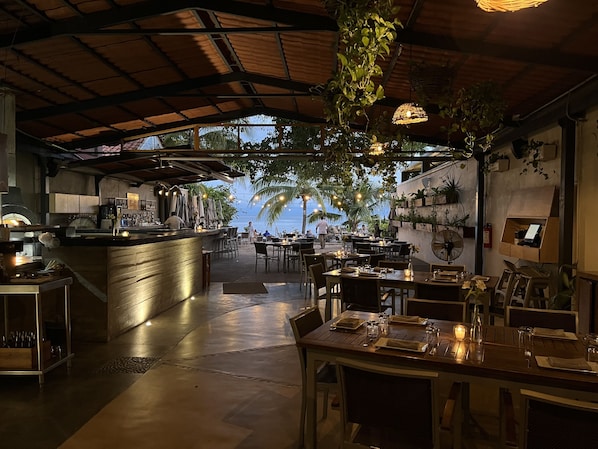 An indoor restaurant with a view of the beach, featuring a bar area, dining tables, and hanging plants.