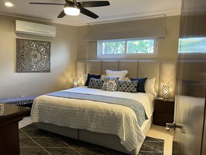 A modern bedroom with a large bed, a ceiling fan, an air conditioning unit, a decorative wall piece, a window with a view, and a nightstand with a lamp. - Punta de Mita Beachfront condo: Amazing Views and Fiber Optic Internet (Punta de Mita)