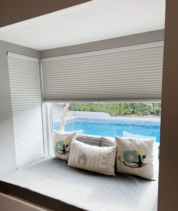 Una ventana en esquina con persianas cerradas, vista a una piscina, y tres almohadas con diseño de pez y la palabra "HOME".