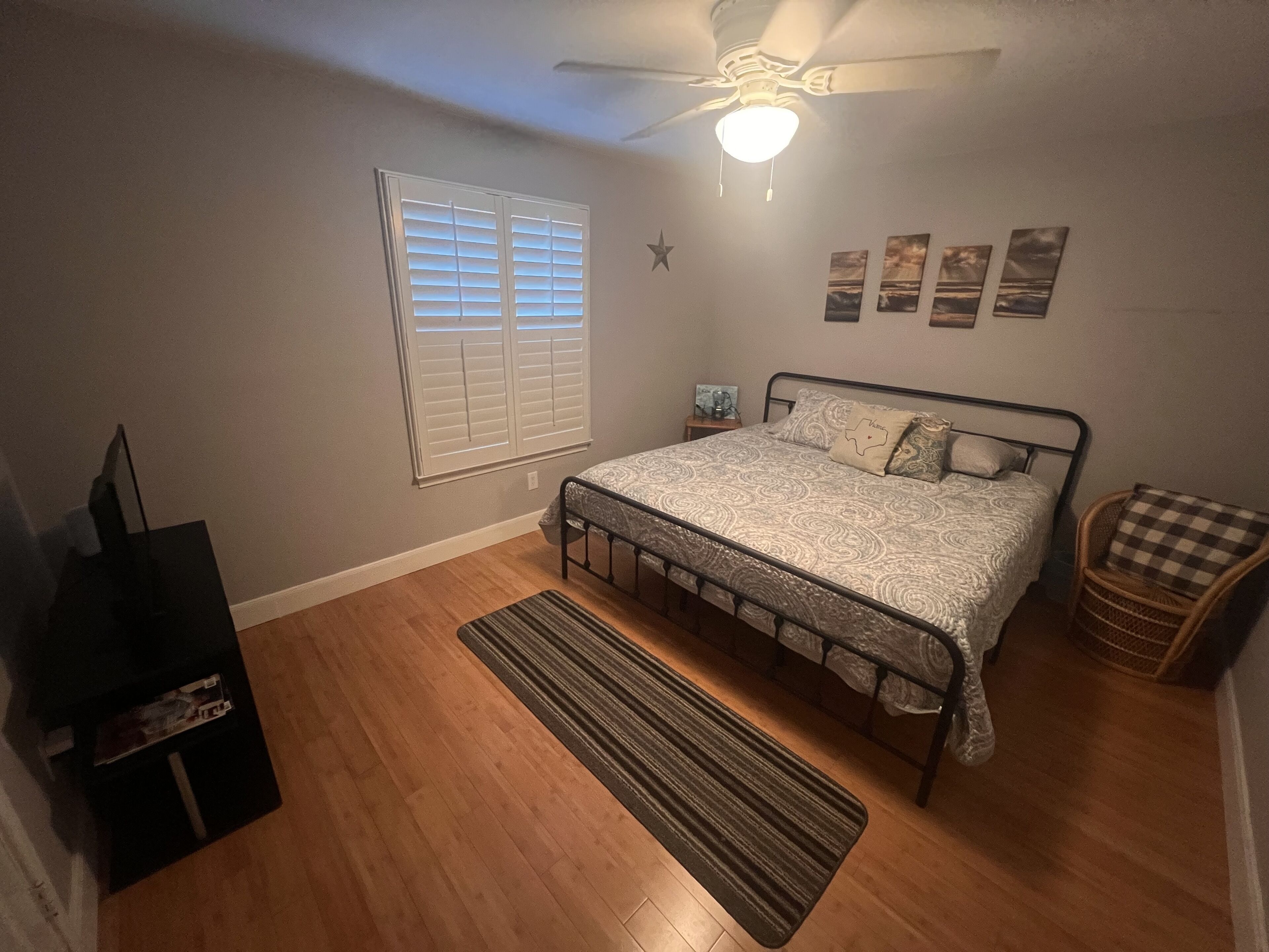 A bedroom with a bed, a ceiling fan, a window with blinds, a small bedside table, and a wall-mounted shelf.