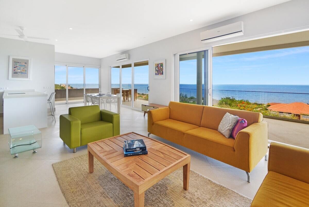 A modern living room with a wooden coffee table, a green armchair, and a mustard sofa. There is a large window offering a view of the sea.