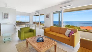 A modern living room with a wooden coffee table, a green armchair, and a mustard sofa. There is a large window offering a view of the sea.