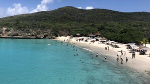 A beach with clear turquoise water, people swimming and walking, and a backdrop of green hills.