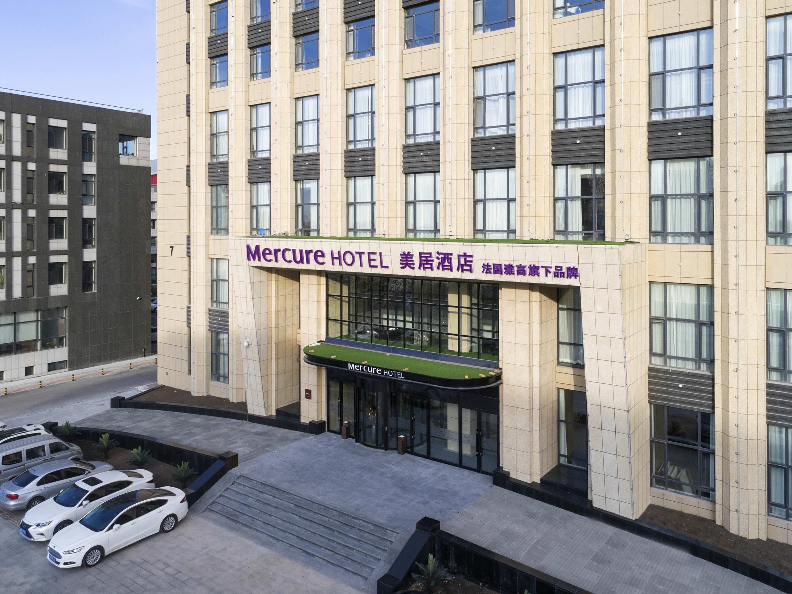 Een hotel met een moderne gevel, meerdere ramen en een groene luifel. Er zijn diverse auto's geparkeerd voor het hotel.