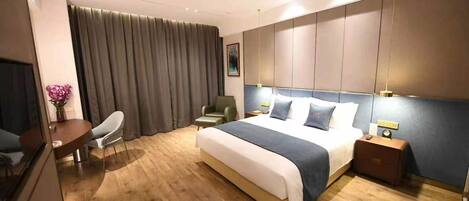 A modern hotel room with a large bed, a desk with a chair, a small table with a vase of flowers, and a large window with curtains.