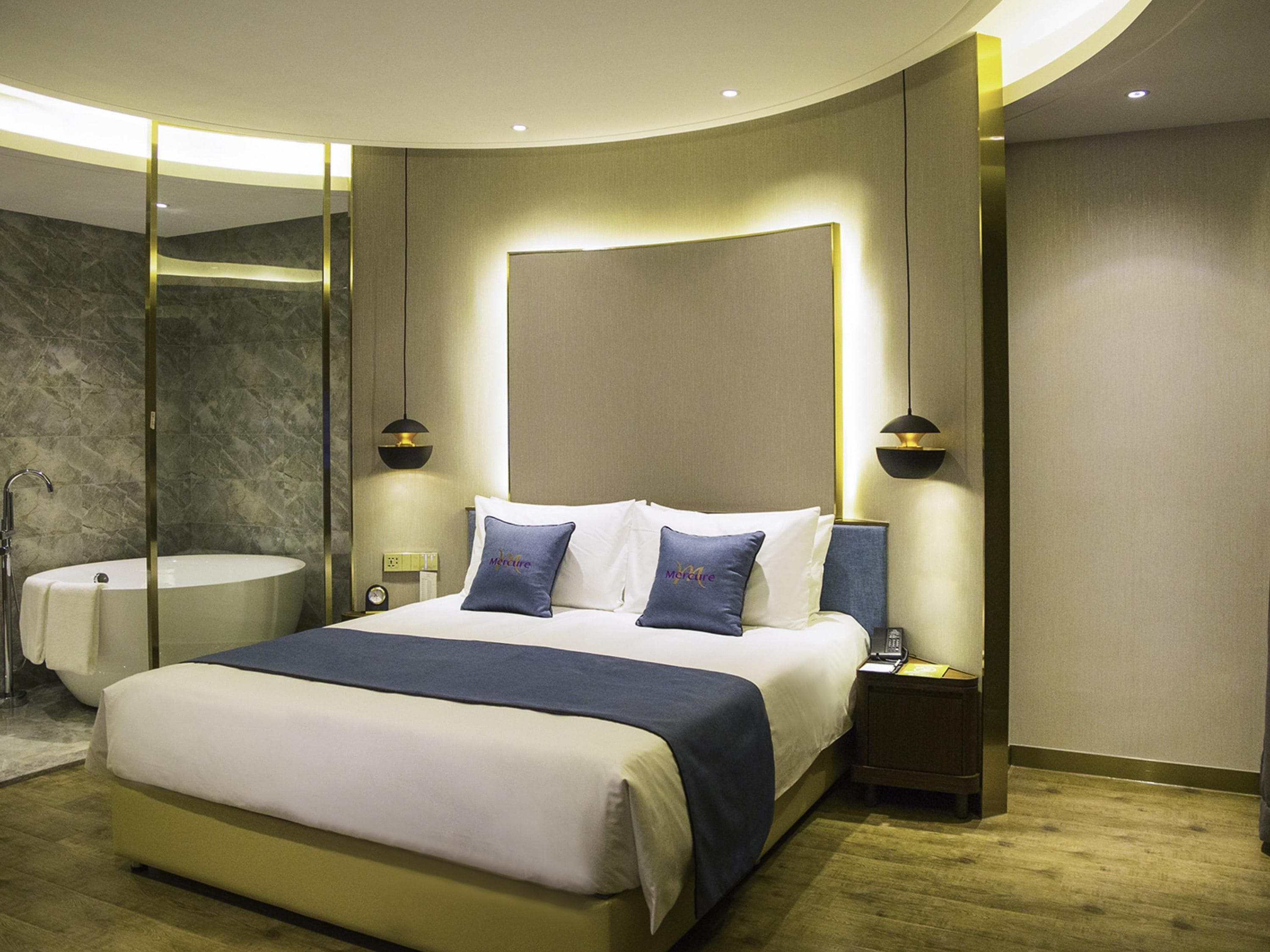 A modern hotel room with a large bed, bedside lamps, a round mirror, and a built-in bathtub.