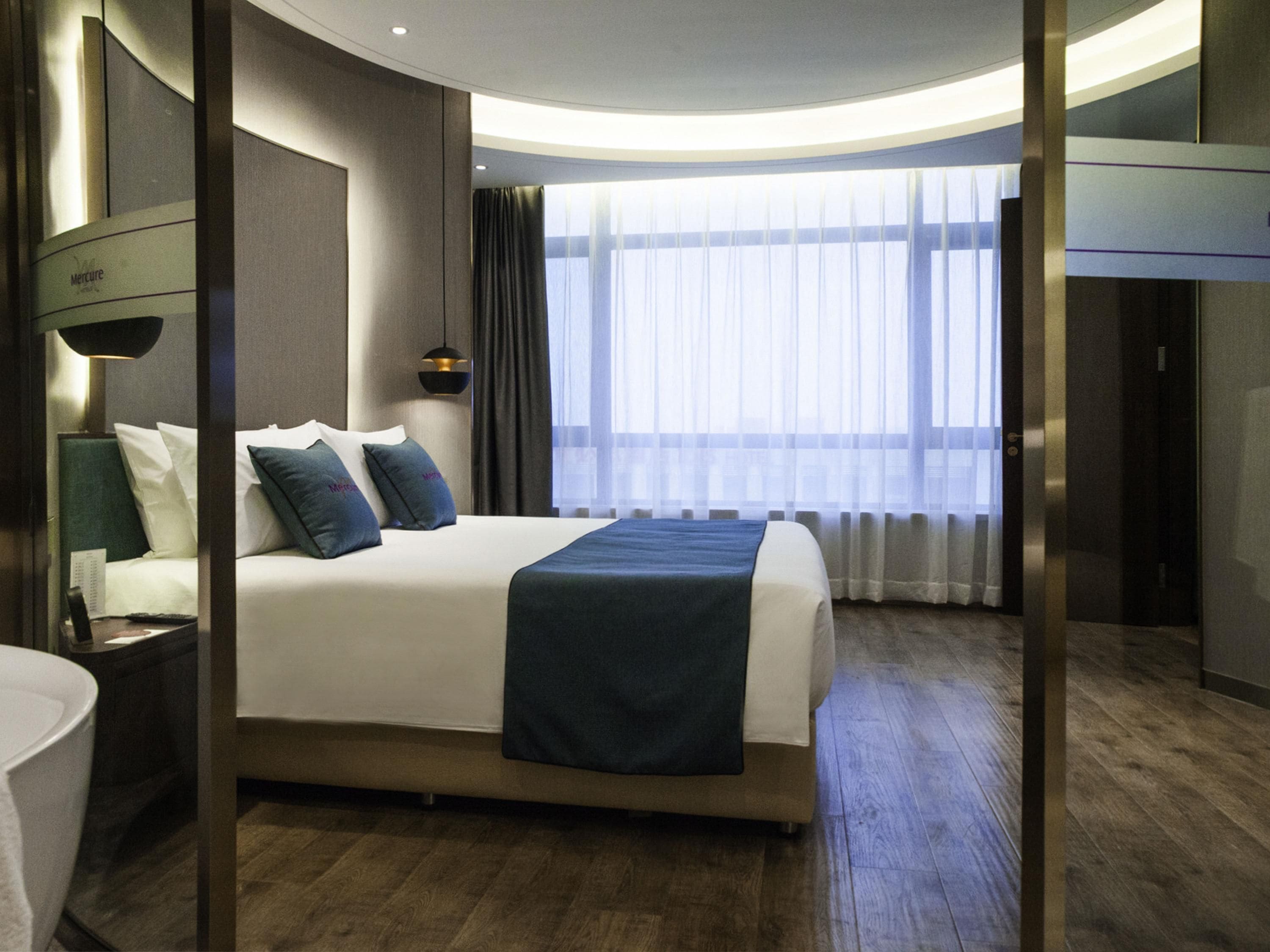 Quarto de hotel moderno com uma cama grande, mesa de cabeceira com abajur, janela com cortinas leves e vista para a cidade.