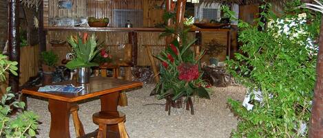 A rustic wooden table with a metal pitcher and a bowl of fruit in a thatched-roof room with bamboo walls and various plants.