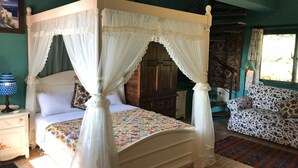 A four-poster canopy bed with a floral bedspread, a nightstand with a lamp, a patterned sofa, and a view of the outdoors through a window.