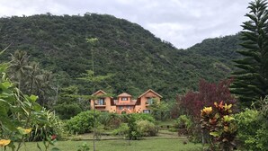 A two-story house with a balcony, surrounded by lush greenery and hills in the background.