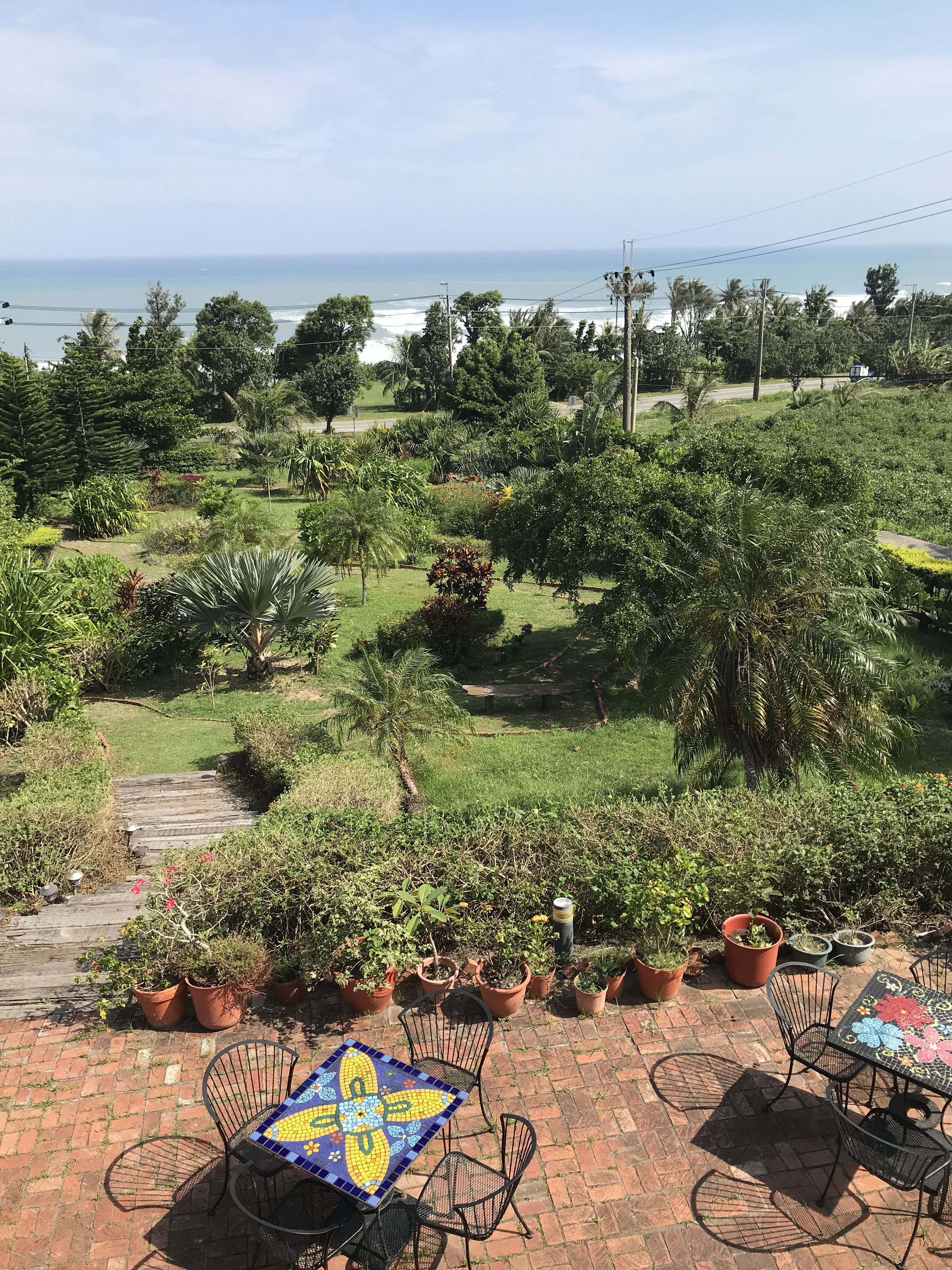 Un patio al aire libre con una mesa y sillas de cerámica, rodeado de plantas en macetas y vistas al océano.